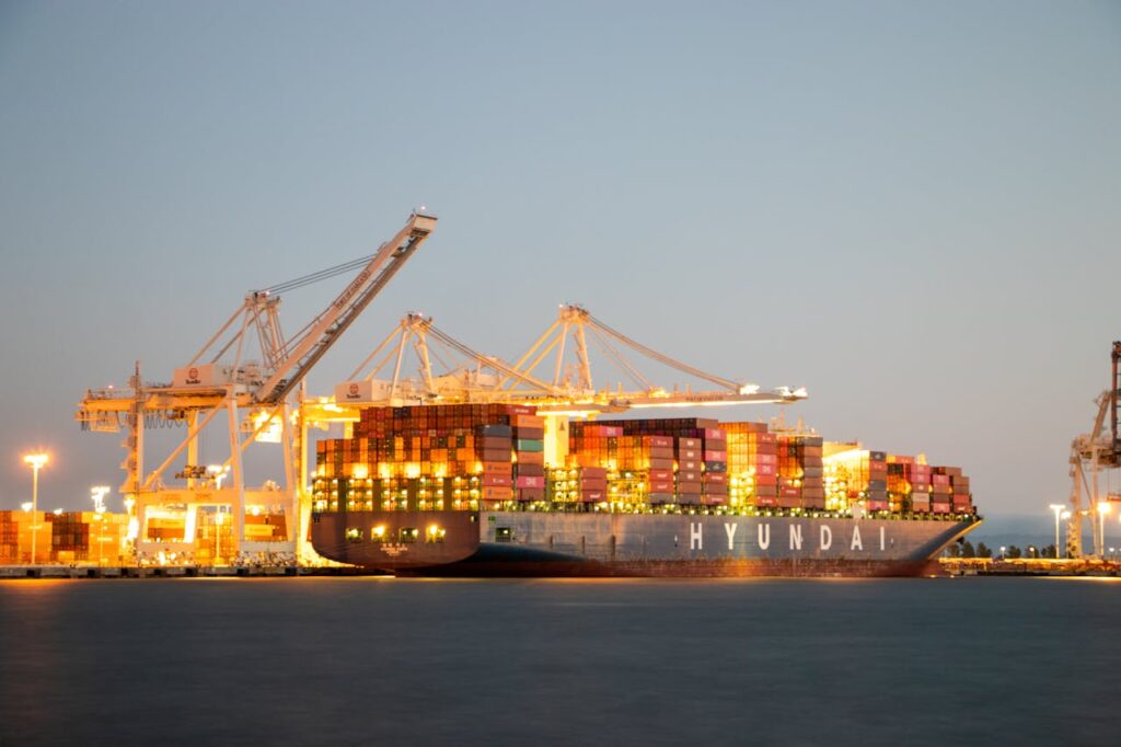 Container ship docked at port under cranes during twilight, illuminated against the evening sky.